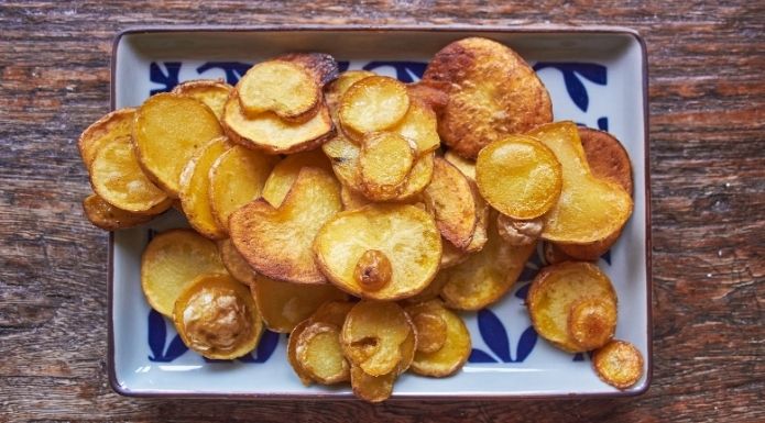 A plate of home-cooked, quarter-inch thick fried potato slices showing uneven browning, illustrating why William Kitchiner's 1817 recipe produces traditional cottage fries rather than modern potato chips.