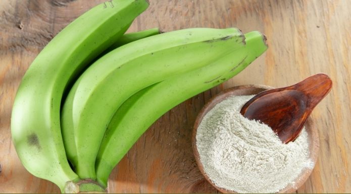 Banana flour in bowl with a bunch of fresh green bananas. 